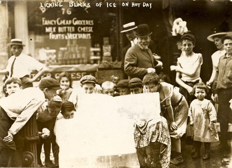 A sepia-toned, historic photograph shows a group of children and a few adults gathered around a large block of ice on the street. Some children are licking the ice. In the background, a grocery store window displays various items. The caption reads, "Licking blocks of ice on hot day.