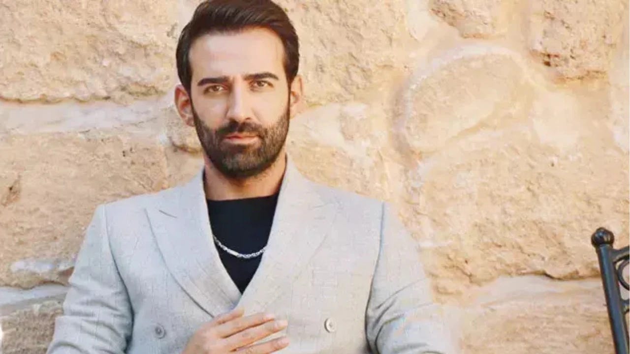 Ferit Kaya, açık gri takım ceket ve siyah tişört kombinini gümüş bir kolyeyle tamamlamış halde taş duvar önünde poz verirken. Bakımlı sakalı ve etkileyici bakışlarıyla profesyonel bir çekim için kameraya bakıyor.