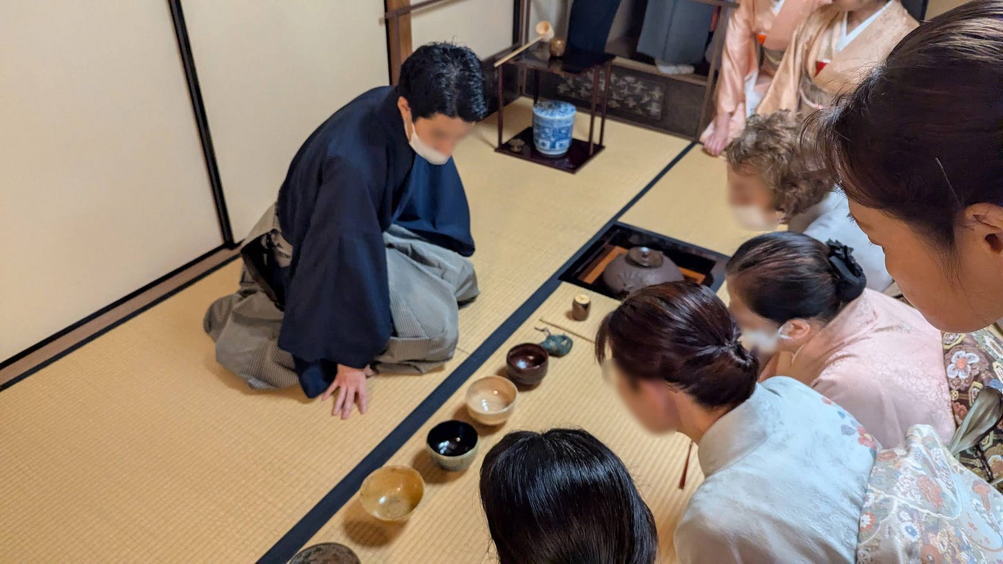 Tea ceremony guests in kimono gather around a young man explaining tea bowls that were used in the ceremony.
