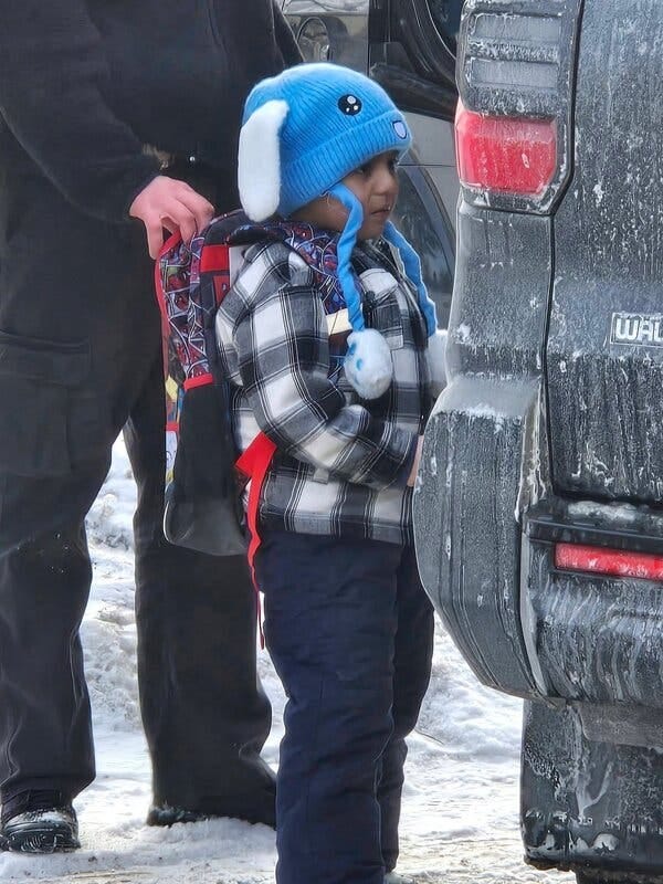 A boy in a blue hat, a coat and a backpack is being led into a vehicle. A boy in a blue hat, a coat and a backpack is being led into a vehicle.