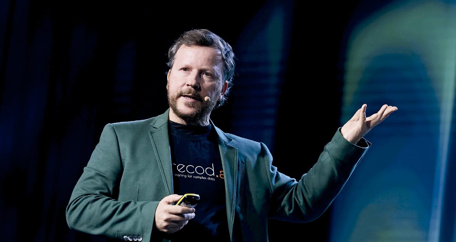 Pessoa em pé sobre palco com iluminação azul e verde ao fundo, usando blazer cinza-esverdeado sobre camiseta preta com texto parcialmente visível, segurando controle remoto na mão esquerda, com braço direito levantado em gesto de apresentação, usando microfone de cabeça, com barba e cabelo grisalho, em postura de orador em ambiente de apresentação ou conferência. Pessoa em pé sobre palco com iluminação azul e verde ao fundo, usando blazer cinza-esverdeado sobre camiseta preta com texto parcialmente visível, segurando controle remoto na mão esquerda, com braço direito levantado em gesto de apresentação, usando microfone de cabeça, com barba e cabelo grisalho, em postura de orador em ambiente de apresentação ou conferência.