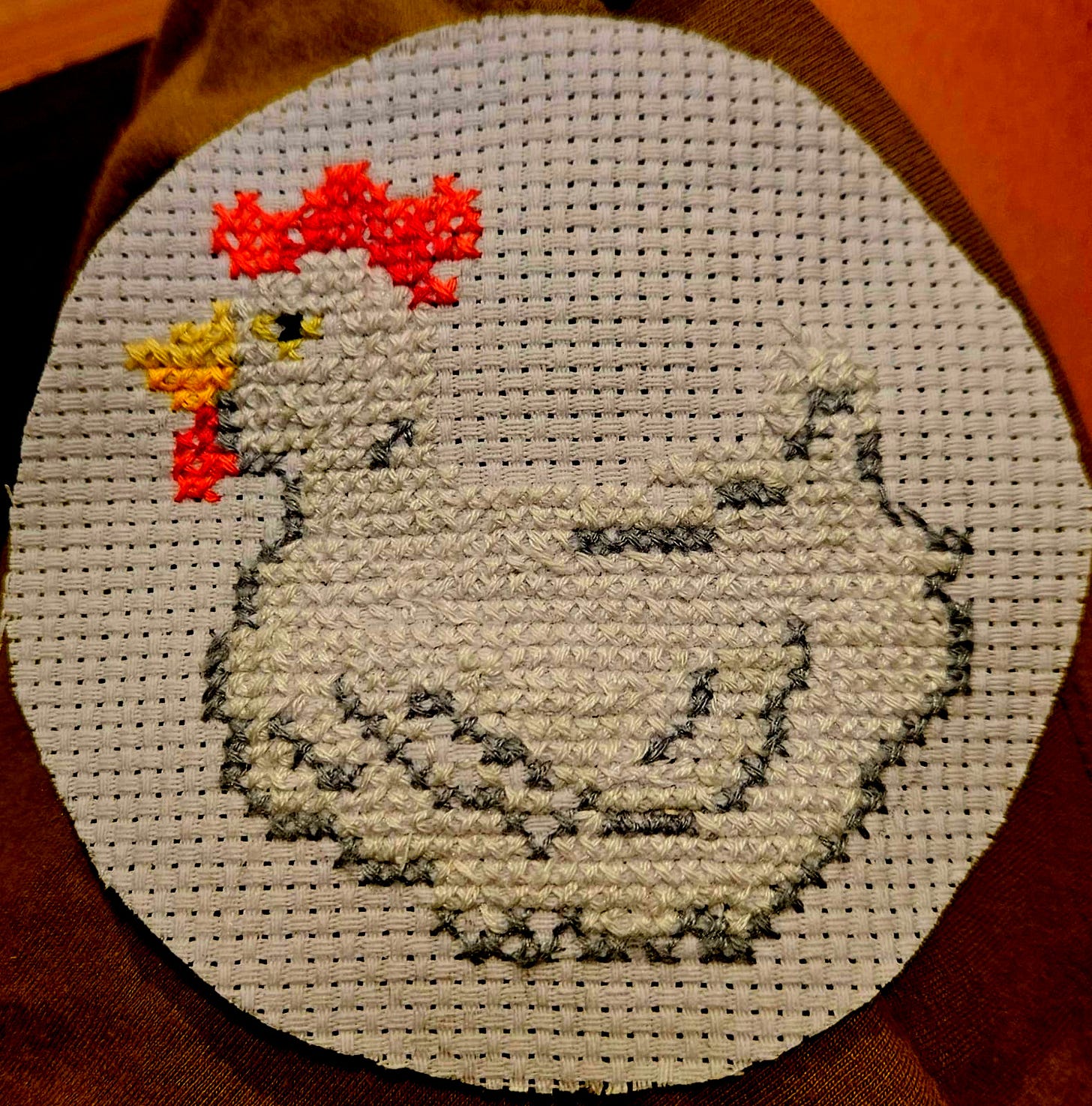 Um cículo de tecido furadinho com uma galinha branca com contornos cinzas bordada em ponto cruz