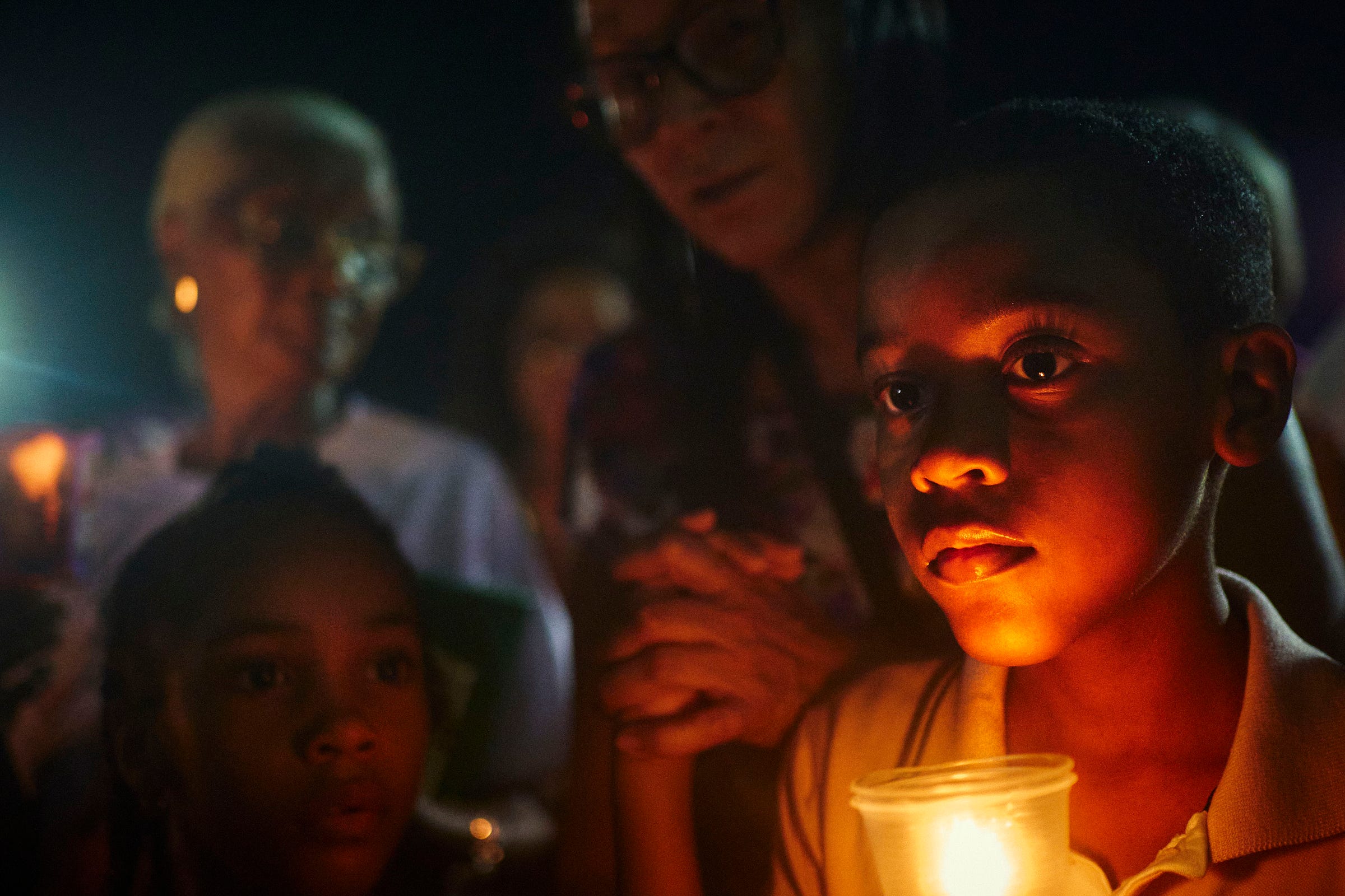 A young boy holds a glowing candle at night, his face illuminated by its light. He is surrounded by others, some with candles, creating a solemn, reflective atmosphere.