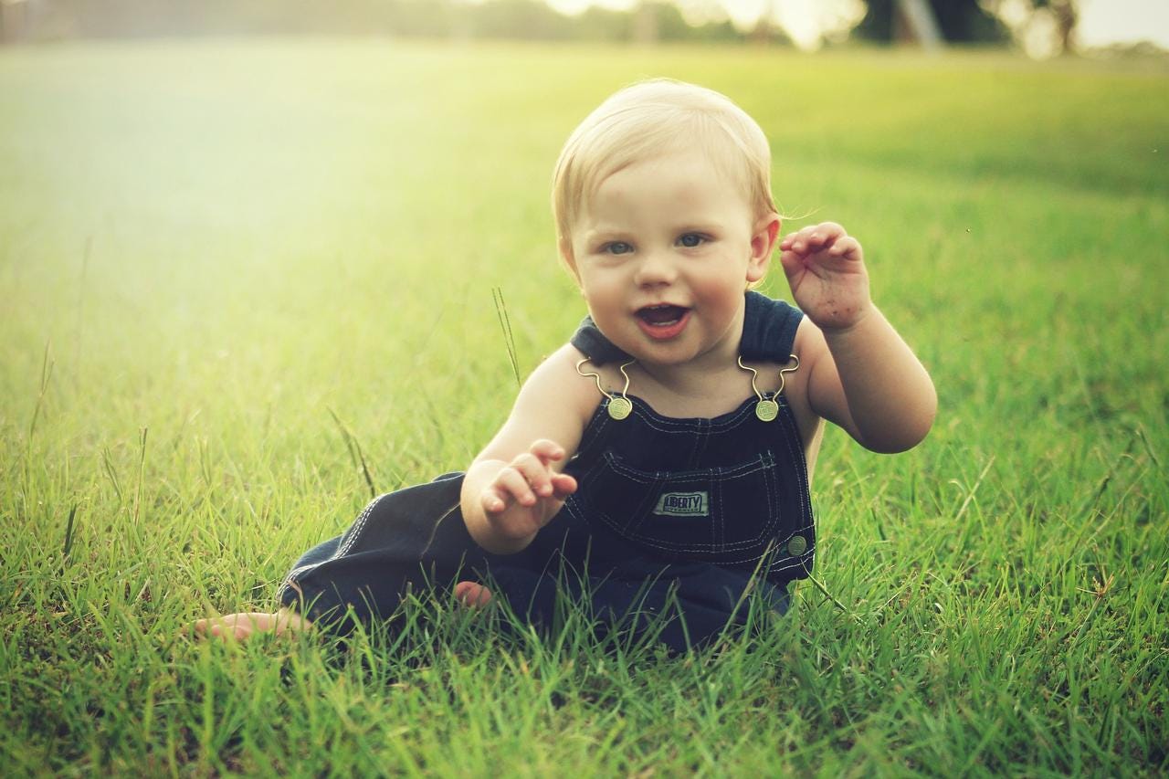 Picture of a cute baby in overalls on a grassy field, lit by a golden sunset.
