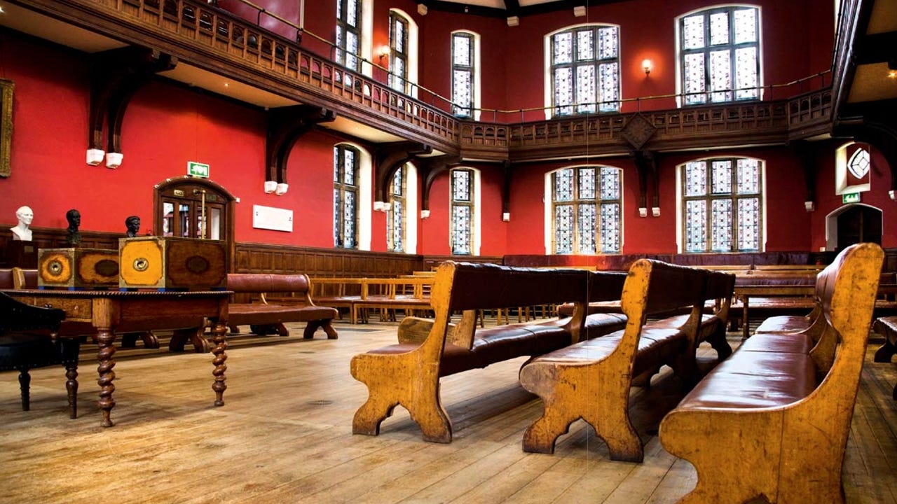 Photograph of the Oxford Union Debating Chamber. The photograph shows the worn wooden floors, wooden benches, red walls and windows with wood galleries on the second floor. Image credit: Barker Evans/Wikimedia Commons (FAL).
