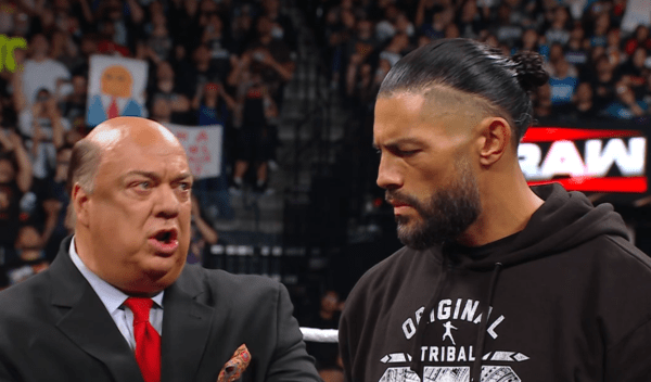 Paul Heyman (homem branco, gordo e calvo) falando algo para Roman Reigns (homem indígena, alto, forte, com barba e cabelos compridos presos num coque.