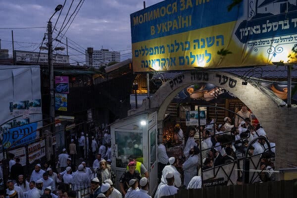 A city street at dusk full of men in skullcaps and white shirts, passing under a brick archway with Hebrew lettering on it.