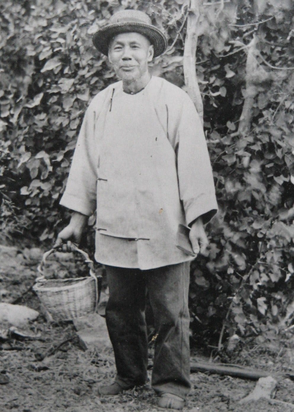 A black and white old photo with a middle-age looking Asian man standing in the field, holding a basket and smiling at camera. A black and white old photo with a middle-age looking Asian man standing in the field, holding a basket and smiling at camera.