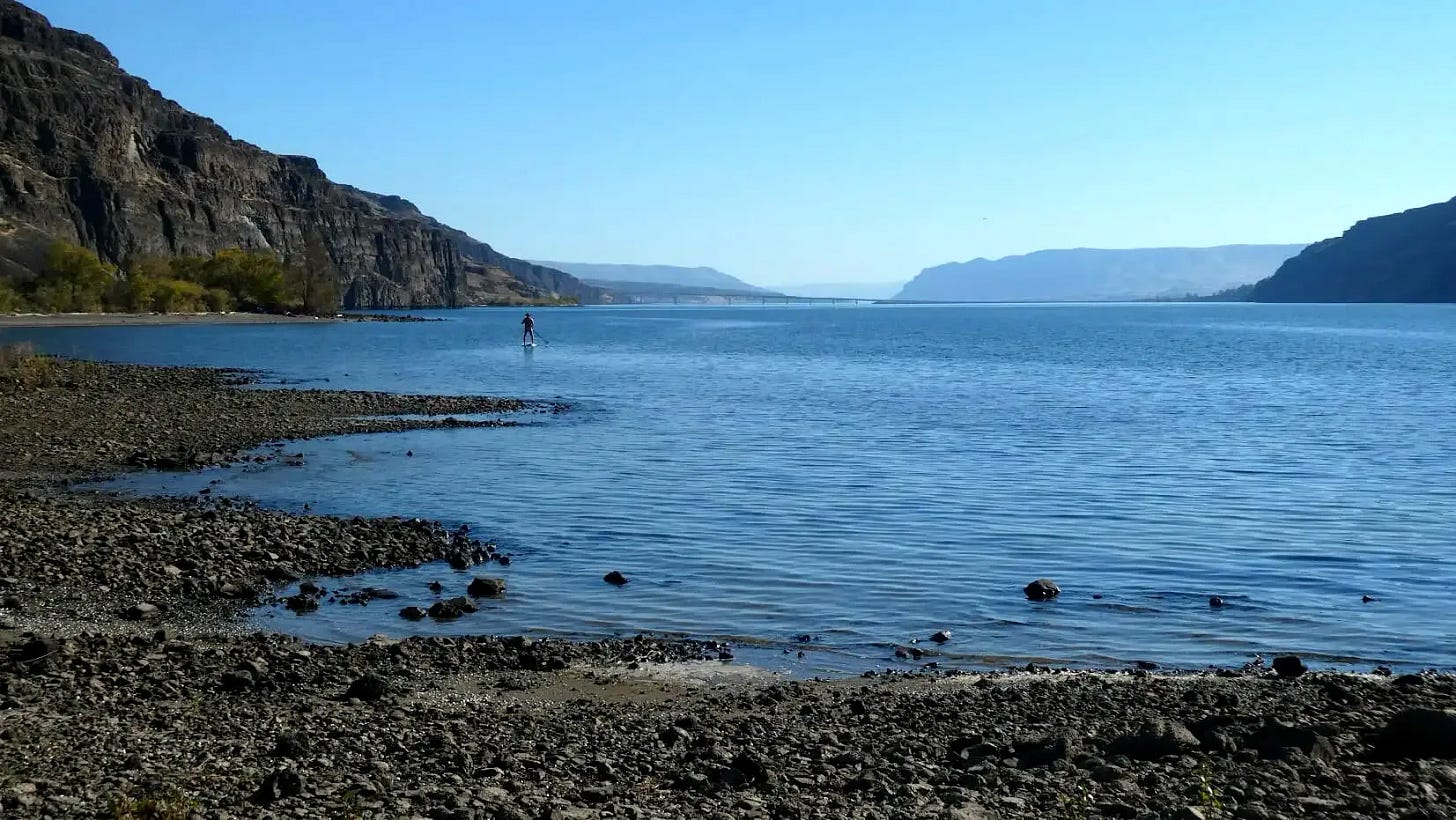 A view of Vantage Beach with someone standing on a paddle board in the water in the deep distance. 
