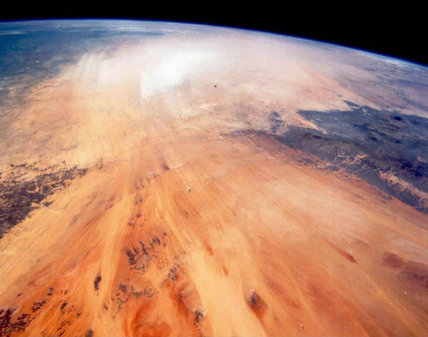 Sand streaks visible in an image taken from space, with orange sand, a cloud in center where sand is carried by the wind, and dark areas corresponding to the Tibesti Mountains and Ennouchi Plateau Sand streaks visible in an image taken from space, with orange sand, a cloud in center where sand is carried by the wind, and dark areas corresponding to the Tibesti Mountains and Ennouchi Plateau