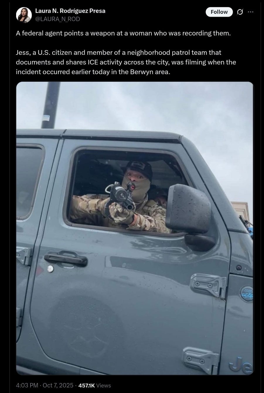 Alt text: A man in camouflage clothing and a face covering sits inside a gray Jeep, pointing a weapon out of the open driver-side window. The gun is aimed directly at the camera. The man wears gloves and a dark cap, and his eyes are visible above the mask. The vehicle’s door and side mirror are wet with raindrops.


