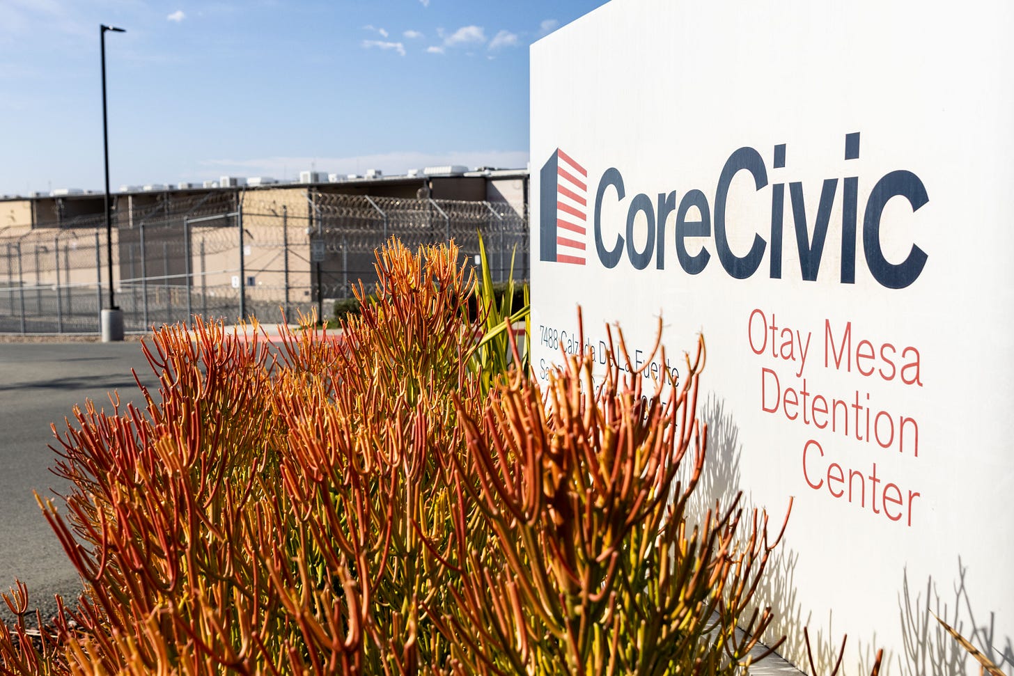A sign that says CoreCivic Otay Mesa Detention Center stands in front of a building with a tall fence and concertina wire