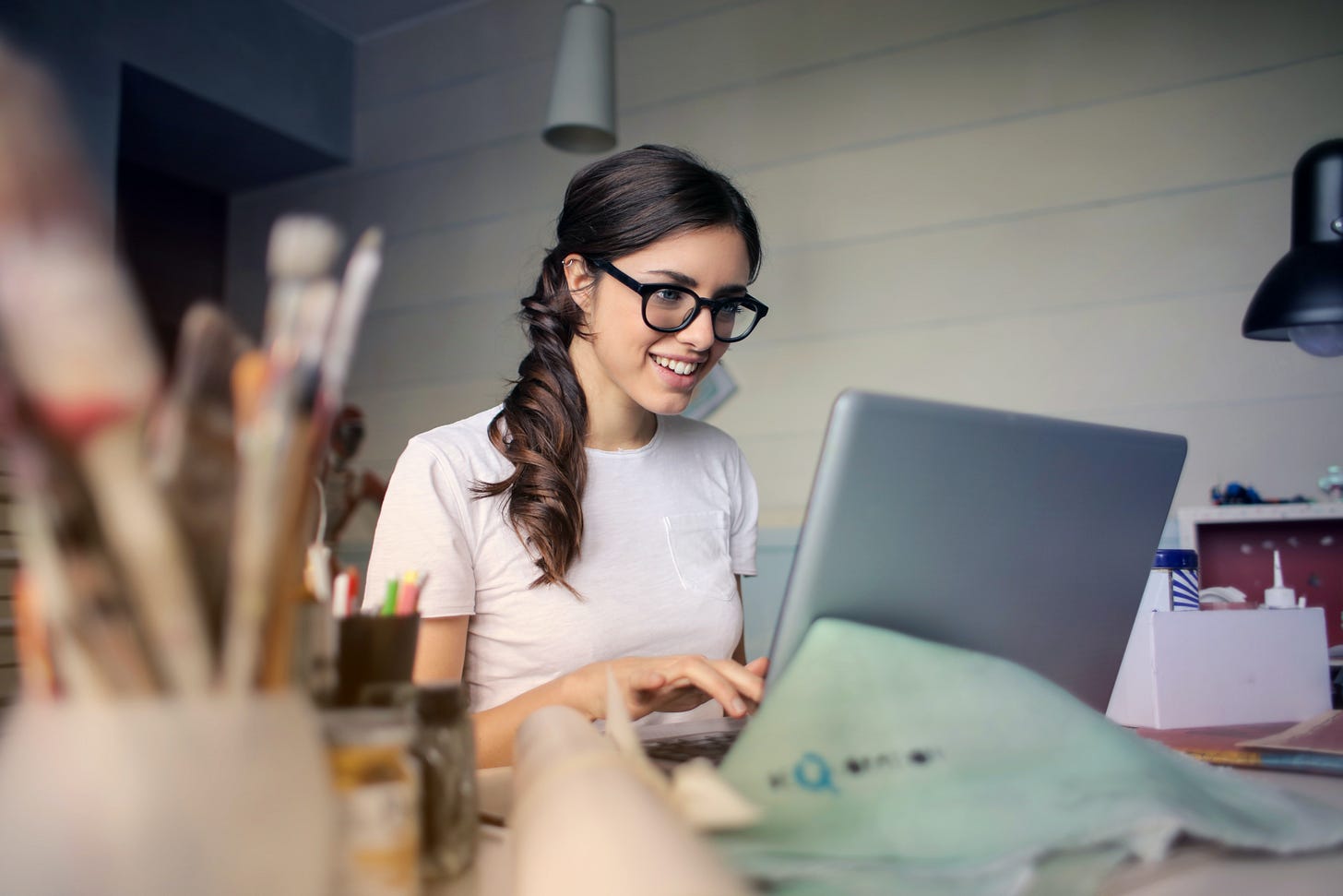 Woman with glasses smiling at laptop, surrounded by art supplies on a desk.