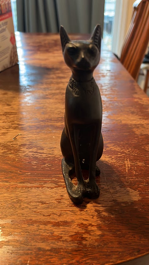 A black figurine of an Egyptian cat. A road lined with blossoming cherry trees. A necklace of aqua colored stones that are cracked and broken. An empty seat and rainy window at a cafe, with coffee on the table. An ikebana arrangement of tall daffodils. A campfire burning inside a potbellied chiminea. A collection of art supplies and a plaster angel. A watercolor sketch of cartoon spiders. A whiteboard at my job bearing the question, What's Your Favorite Thing About Baltimore In The Summer? With many many answers.