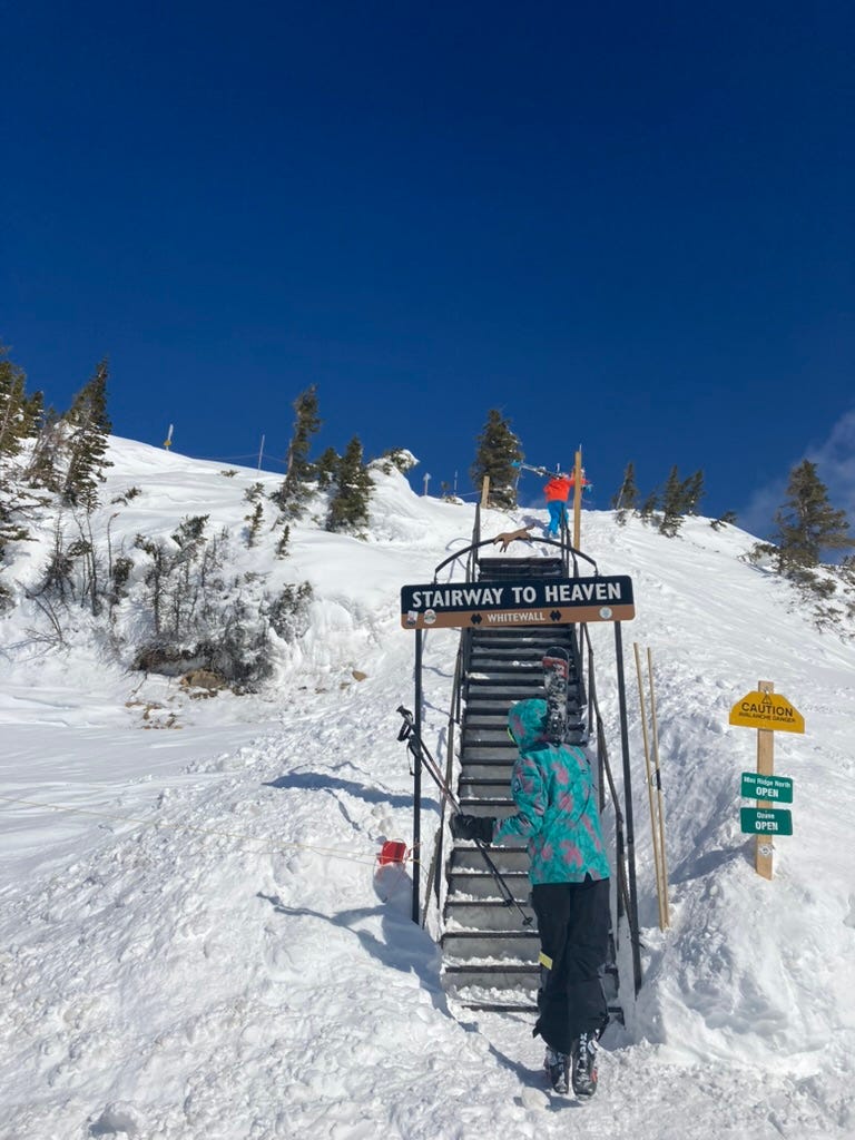 two boys hiking up a set of stairs on a ski hill. A sign overhead says "Stairway to Heaven"