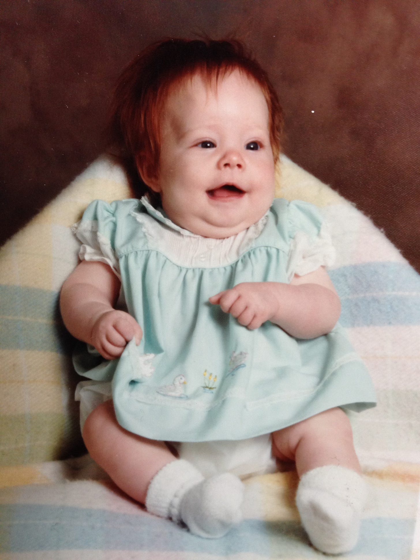 A chubby white baby with red hair and blue eyes wears white socks, a blue dress, and sits on a blanket.