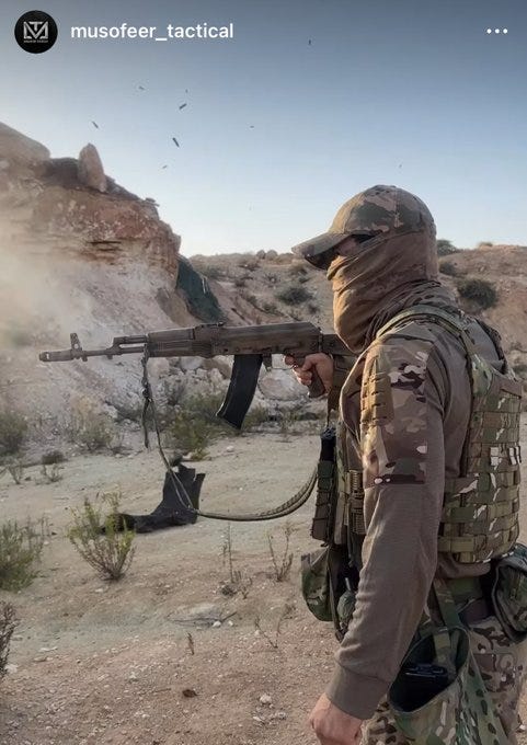 Masked individual in camouflage cap and tactical vest holds AK-74M rifle with sling and magazine in rocky desert landscape under clear sky birds visible in background gear includes backpack and boots.