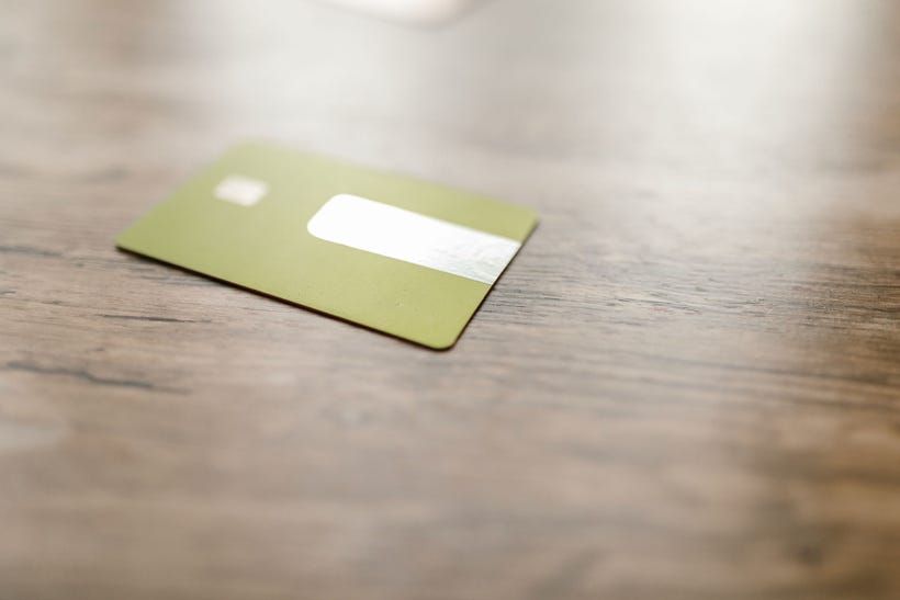 A blurred expanse of wood grain. The center horizontal strip of it is in focus, along with a blank gold-tone credit card lying slightly off to the left. It has no identifying markings, just a shiny strip and the blurred shadow of a chip.