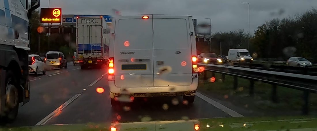 A landscape colour photo. Central in the image is a white van. A Ford Transit. It has its rear lights on. The photo is taken through a car windscreen on a rainy evening. So some of the lights are distorted. This is the van from the subtitle. It has it's brakes on, but the fog lights and the reversing lights are the ones illuminated. This is a photo taken from dashcam footage.