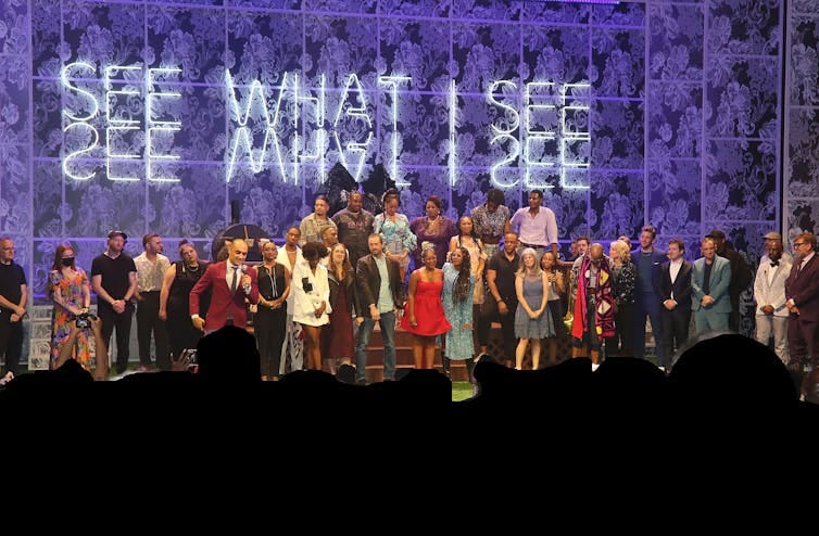 A large crowd of people onstage with a sign behind them that reads 'See What I See'