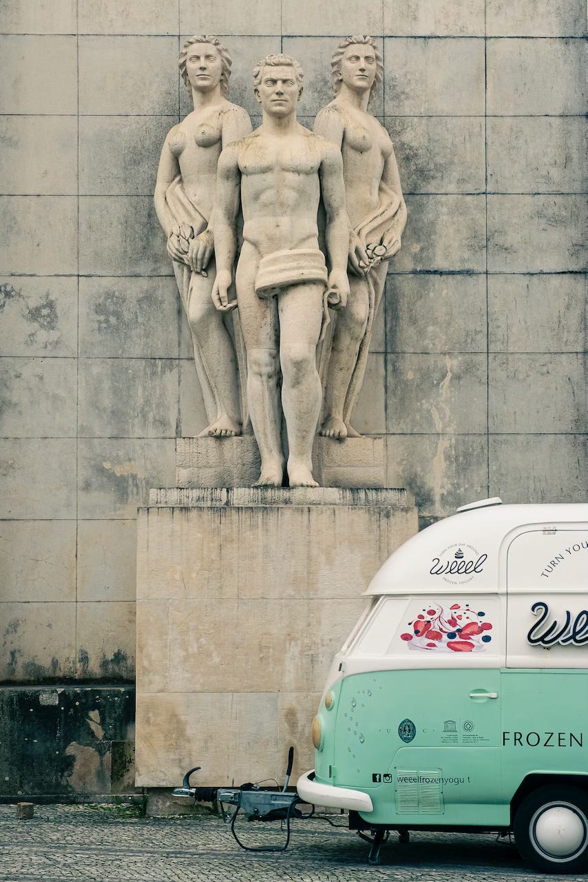 A 1950's three-figure statue is seen beyond a pale green frozen yogurt food truck, on the square in front of the old university building, in Coimbra.