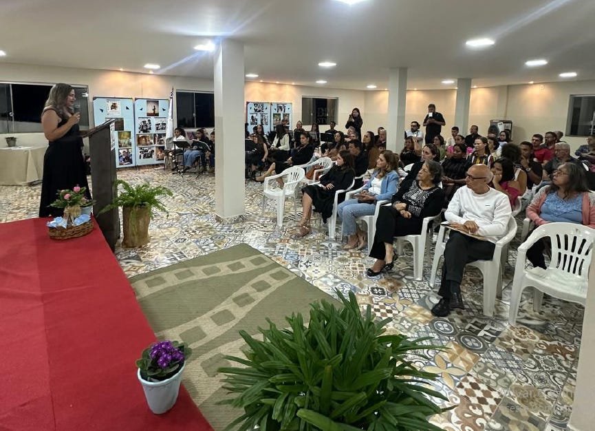 #descrevemospravocê  A imagem mostra um evento realizado em ambiente fechado, com piso de azulejos coloridos e iluminação clara. À esquerda, uma mulher de vestido preto fala ao microfone atrás de um púlpito, sobre um palco coberto com tapete vermelho e decorado com plantas e vasos de flores. O púlpito tem ao lado um cesto com enfeites e um vaso com samambaia.  À frente da mulher, o público está sentado em cadeiras brancas de plástico. As pessoas estão atentas, ouvindo a fala, com expressões concentradas e respeitosas. Entre os presentes, há homens e mulheres de diferentes idades, vestindo roupas casuais e elegantes. Algumas pessoas seguram cadernos ou papéis, indicando um possível caráter formativo ou de homenagem do evento.  Ao fundo, há painéis expositivos com fotografias e imagens, organizadas sobre suportes brancos que cobrem parte da parede. Em uma das janelas, vê-se a bandeira do Brasil. Ao lado da plateia, uma mulher em cadeira de rodas também acompanha a atividade. O espaço é amplo, com colunas brancas e teto baixo, iluminado por luzes redondas embutidas.  A atmosfera é acolhedora e organizada, sugerindo um encontro cultural, educativo ou comemorativo.
