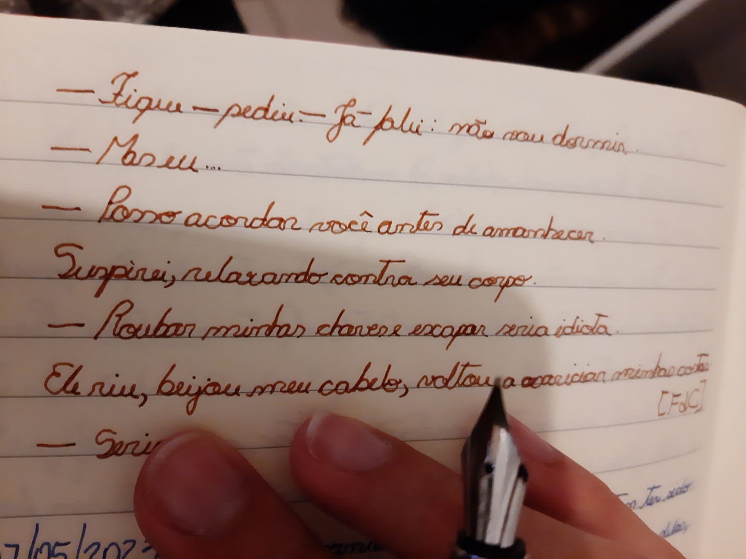 Foto de um quarto de página de um caderno pautado, com texto escrito em tinta marrom à mão. "Fique," pediu. "Já falei: não vou dormir." Parágrafo. "mas eu..." Parágrafo. "Posso acordar você antes de amanhecer." Parágrafo. Suspirei, relaxando contra seu corpo. Parágrafo. "Roubas minhas chaves e escapar seria idiota. Parágrafo. "Ele riu, beijou meu cabelo, voltou a acariciar minhas costas." Parágrafo. "Seria." Abaixo, as pontas de meus dedos segurando a caneta-tinteiro, cobrindo algo escrito em tinta azul. Só dá para ler a data: 7 de maio de 2023.
