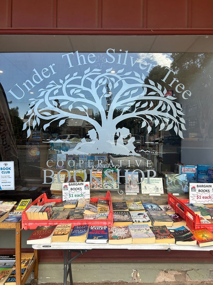 Photos of Bookshops. 1) The window display of Under the Silver Tree Cooperative Bookshop with trolleys of books ready to sell out the front. 2) The outside of The Broken Hill Bookshop - the front of the shop is painted white with a large sign displaying the store's name, the door is open to welcome customers inside. 3) A piece of craft paper on display inside Big Sky Stories divided into two sections. The first says "Is..." and the second says "Needs..." customers have written underneath the headings. 3) A photo of the inside of Big Sky Stories, there are lots of books on display, the inside is bright and welcoming.
