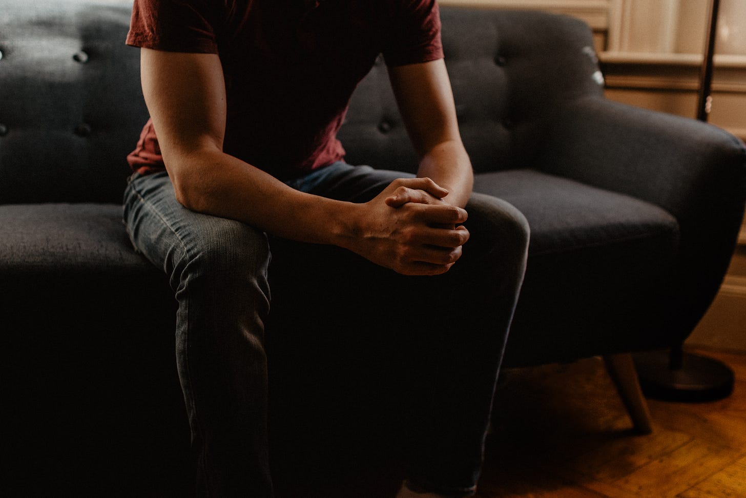 A man sits with hands clenched on a grey couch A man sits with hands clenched on a grey couch