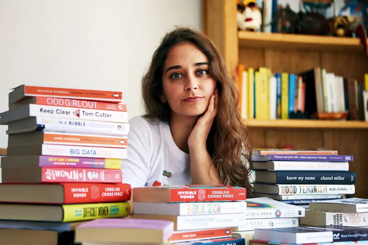 Una donna con i capelli lunghi e mossi è seduta dietro una scrivania sommersa da pile di libri. Guarda l'obiettivo appoggiando il viso a una mano, con un'espressione serena. I titoli dei libri spaziano tra temi di femminismo, sociologia e neurodiversità. Una donna con i capelli lunghi e mossi è seduta dietro una scrivania sommersa da pile di libri. Guarda l'obiettivo appoggiando il viso a una mano, con un'espressione serena. I titoli dei libri spaziano tra temi di femminismo, sociologia e neurodiversità.