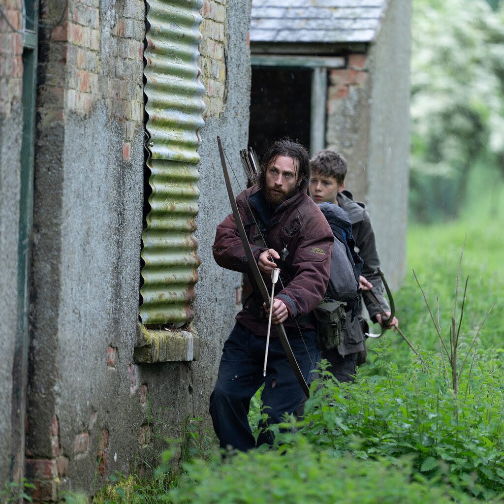 A man and a boy with bows and arrows skulk behind a concrete house.