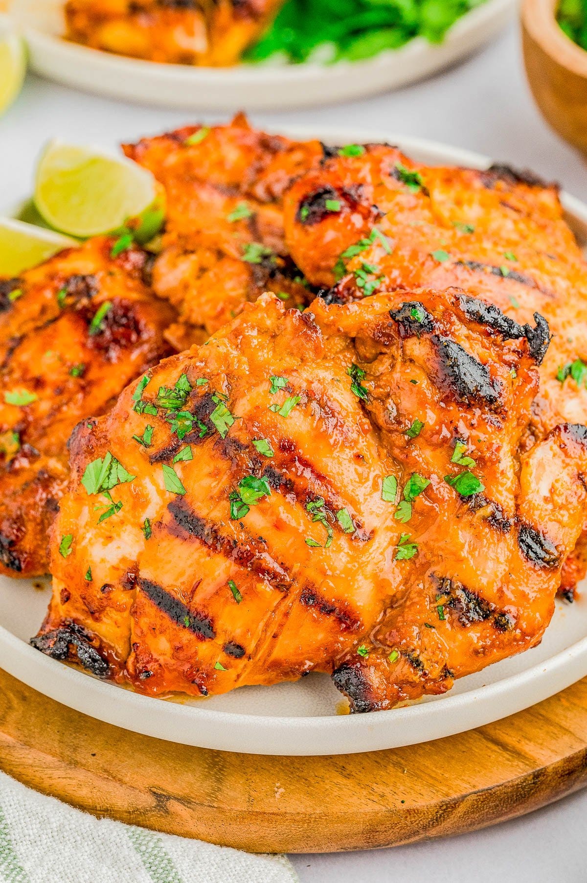 Grilled chicken thighs with a reddish-orange marinade, garnished with chopped herbs, served on a white plate with lime wedges in the background.