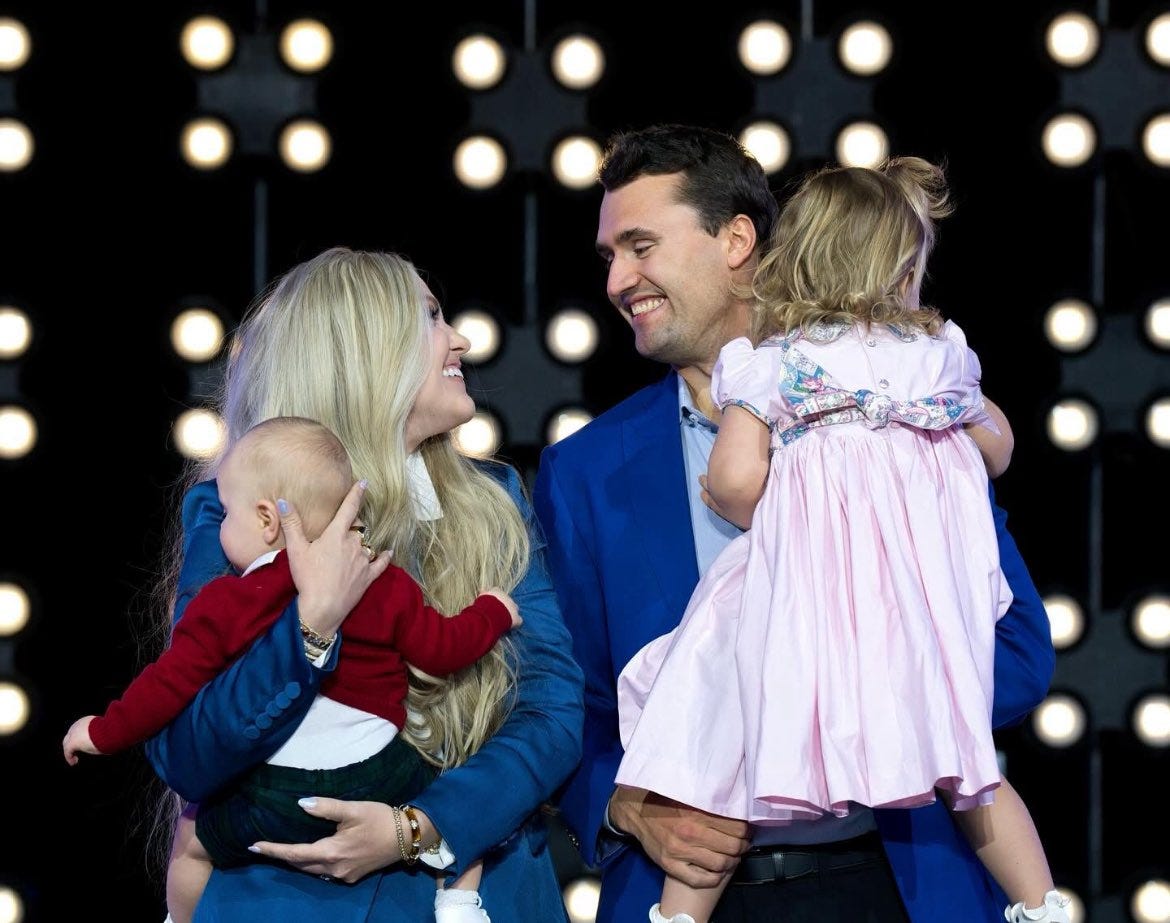 Charlie Kirk stands holding a young girl with blonde hair wearing a pink dress. A woman with long blonde hair holds a baby dressed in a red outfit. Both adults wear blue jackets, standing in front of a backdrop with bright circular lights.