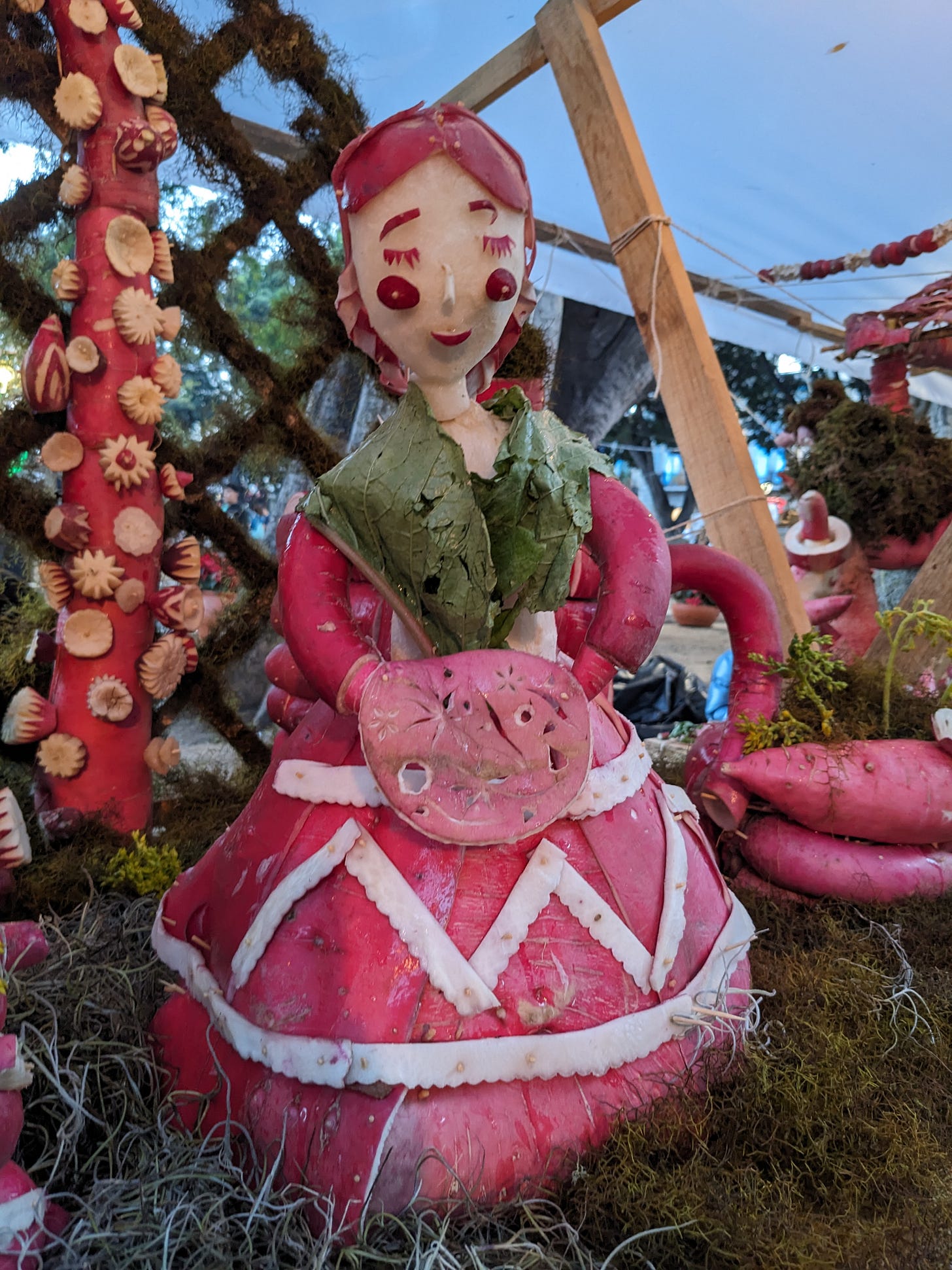 Woman wearing a red dress and green “leaf” shall holding a pastry is roughly carved out of red and white radishes with a background of greens.