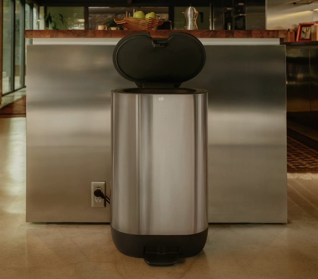 A picture showing the end of a stainless steel kitchen counter with an open Mill food recycler bin, also in stainless steel, looking like a garbage can but blending nicely into its surrounding.