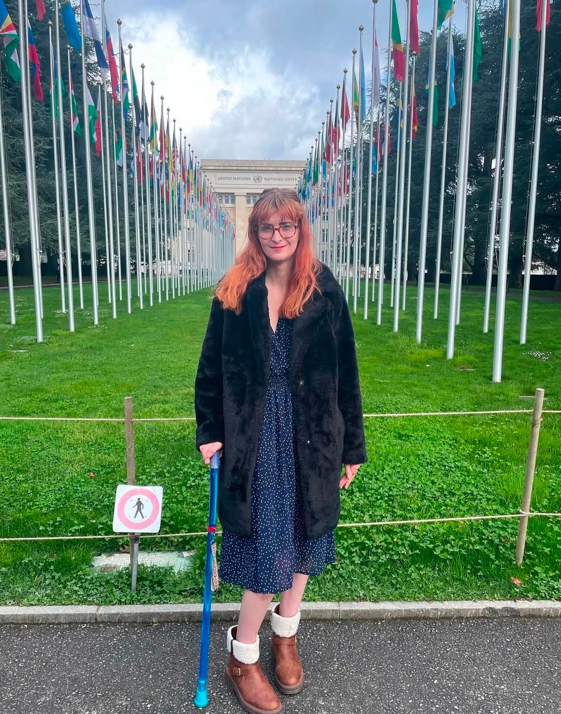 A white person with red hair is standing in front of a green field with flags of lots of different countries. They are wearing a black coat and a blue dress and holding a blue stick which is a mobility aid.