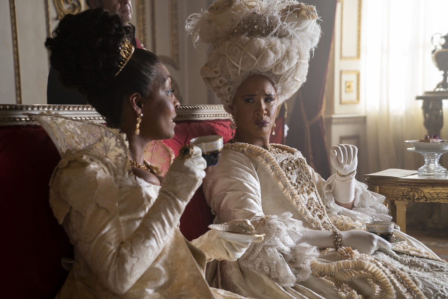 Photo of two older black women in regency era dresses sitting and having tea. They are playing Lady Danbury and Queen Charlotte, two characters who are close friends in Bridgerton Photo of two older black women in regency era dresses sitting and having tea. They are playing Lady Danbury and Queen Charlotte, two characters who are close friends in Bridgerton