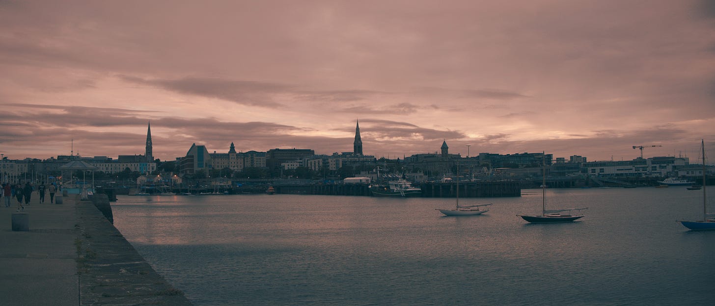 Panoramic view of Dún Laoghaire harbour at dusk, warm pink and mauve clouds overhead, sailboats moored in calm water, and the town's church spires silhouetted along the shoreline.