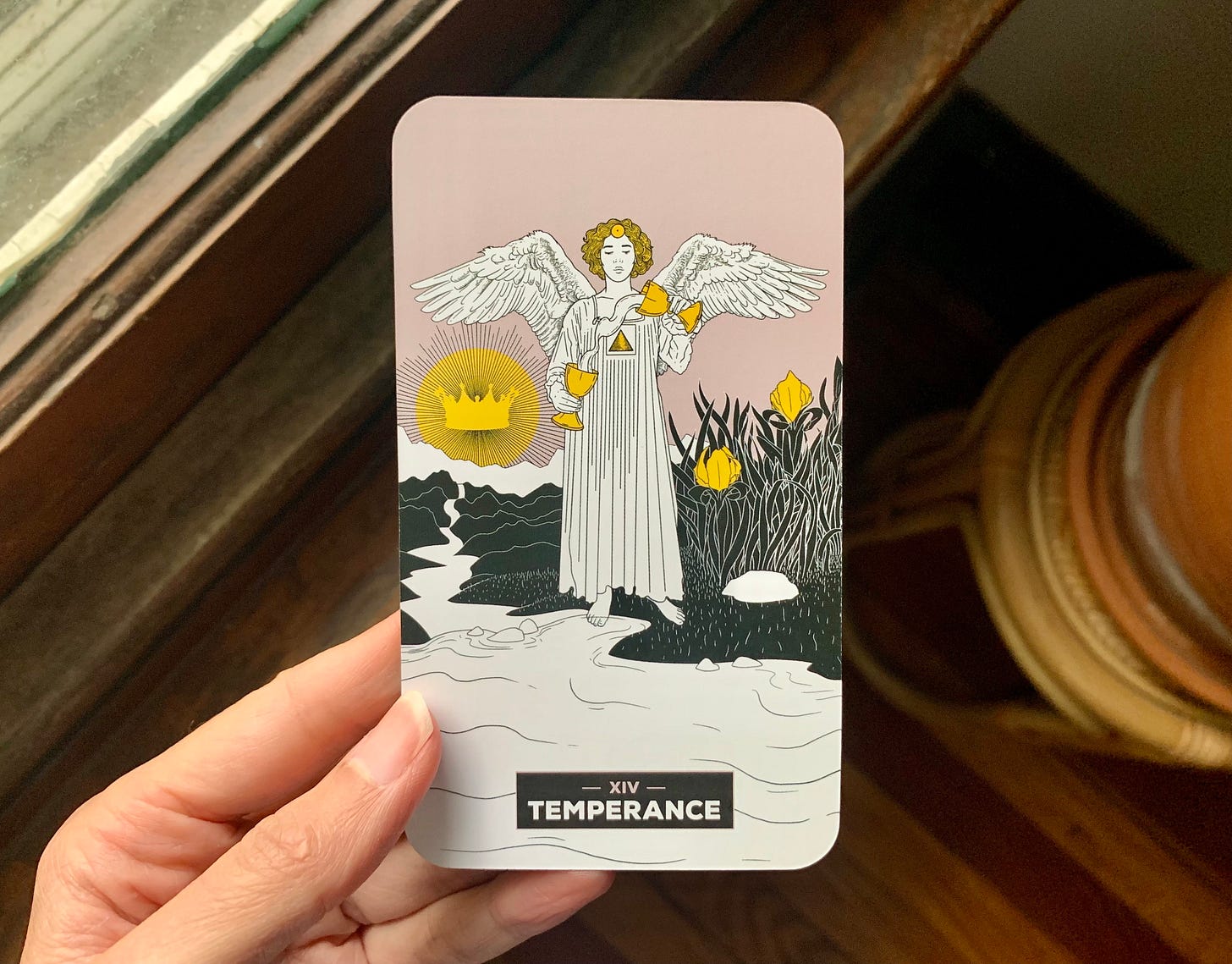 A hand is holding a tarot card, Temperance by Xaviera Lopez for The Change Tarot. In teh image, an angel is holding two cups and water is flowing between them. There is a sun behind them with a crown in it over mountains with a river running through it. There are crocuses. A hand is holding a tarot card, Temperance by Xaviera Lopez for The Change Tarot. In teh image, an angel is holding two cups and water is flowing between them. There is a sun behind them with a crown in it over mountains with a river running through it. There are crocuses.