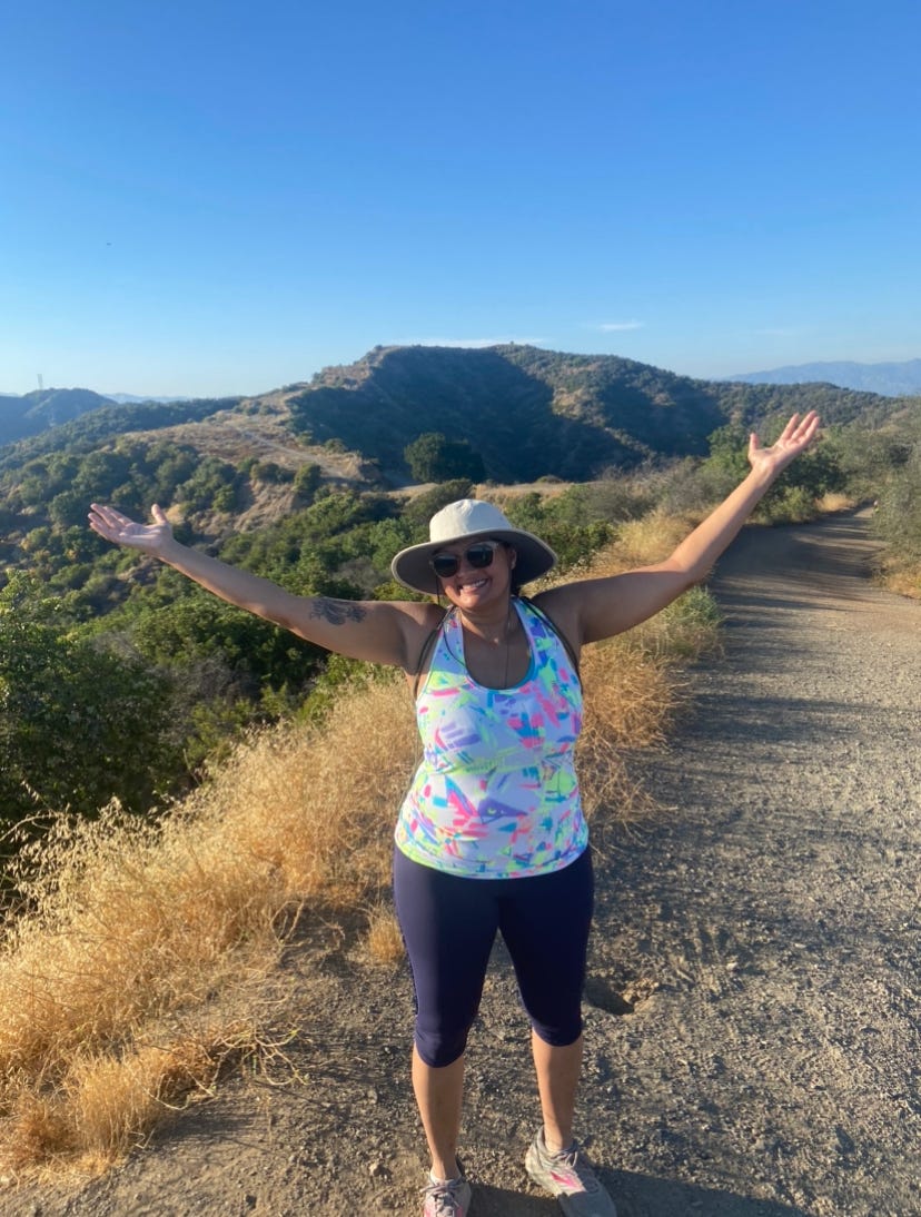 Nisha standing on a hiking trail with her arms up with the air. She is smiling. There is a blue sky above her.