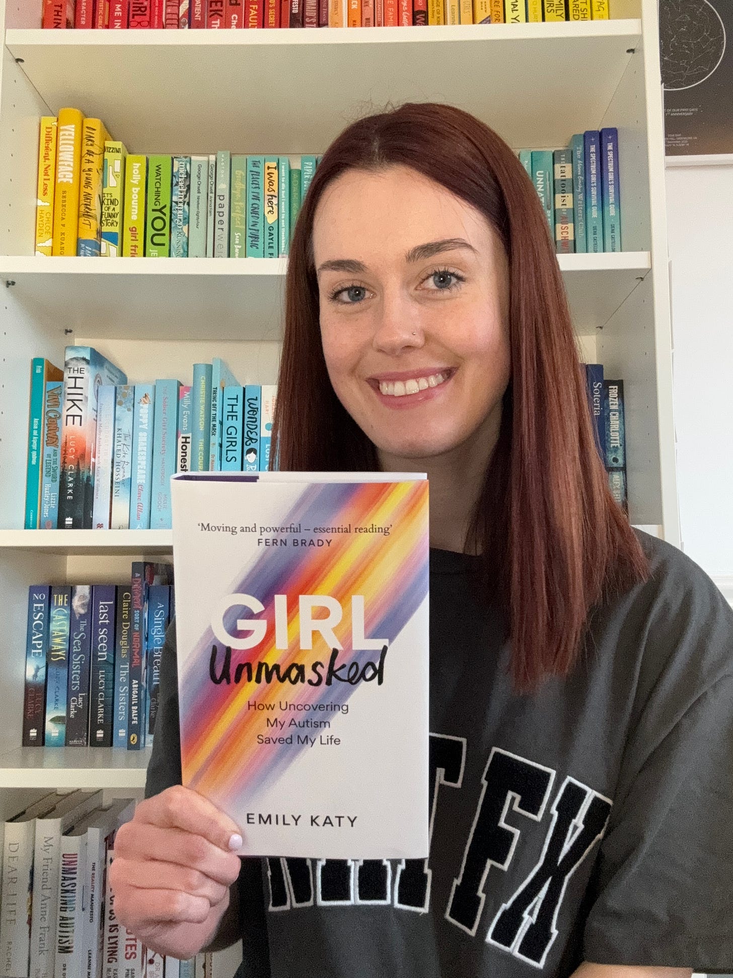 Emily, a young white woman with short reddy brown hair and wearing a White Fox t-shirt is smiling at the camera and holding up her book 'Girl Unmasked: How Uncovering My Autism Saved My Life'. Behind her is her bookshelf where the books are ordered in rainbow colour, with red books at the top and white books at the bottom. 