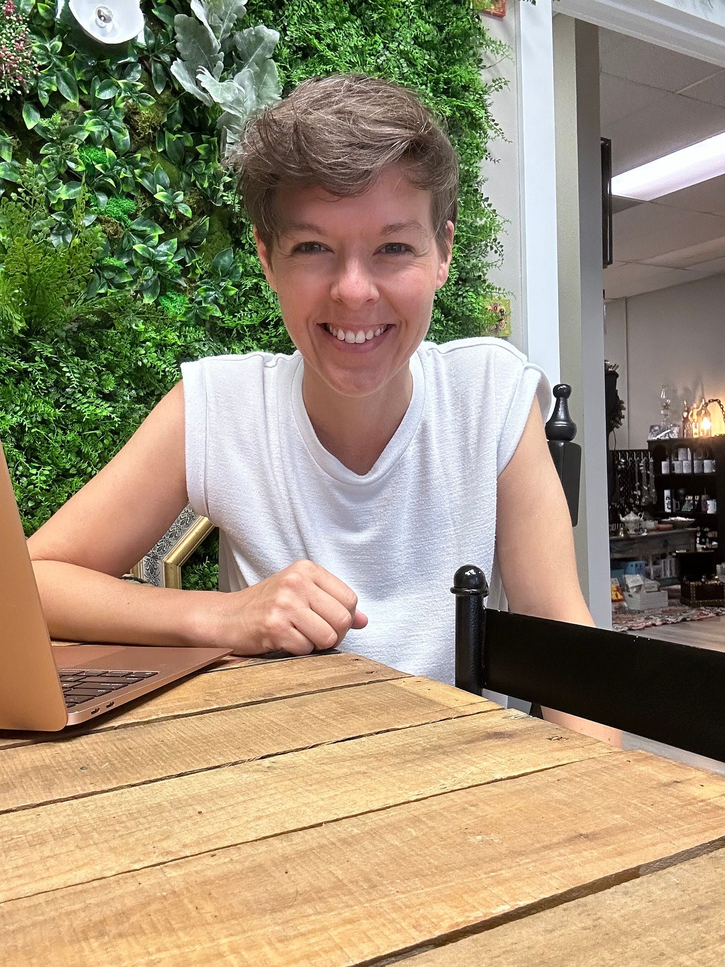 Author Jillian Abby in a white tank top with short brown hair, sitting next to her laptop computer, in front of a faux foliage wall.