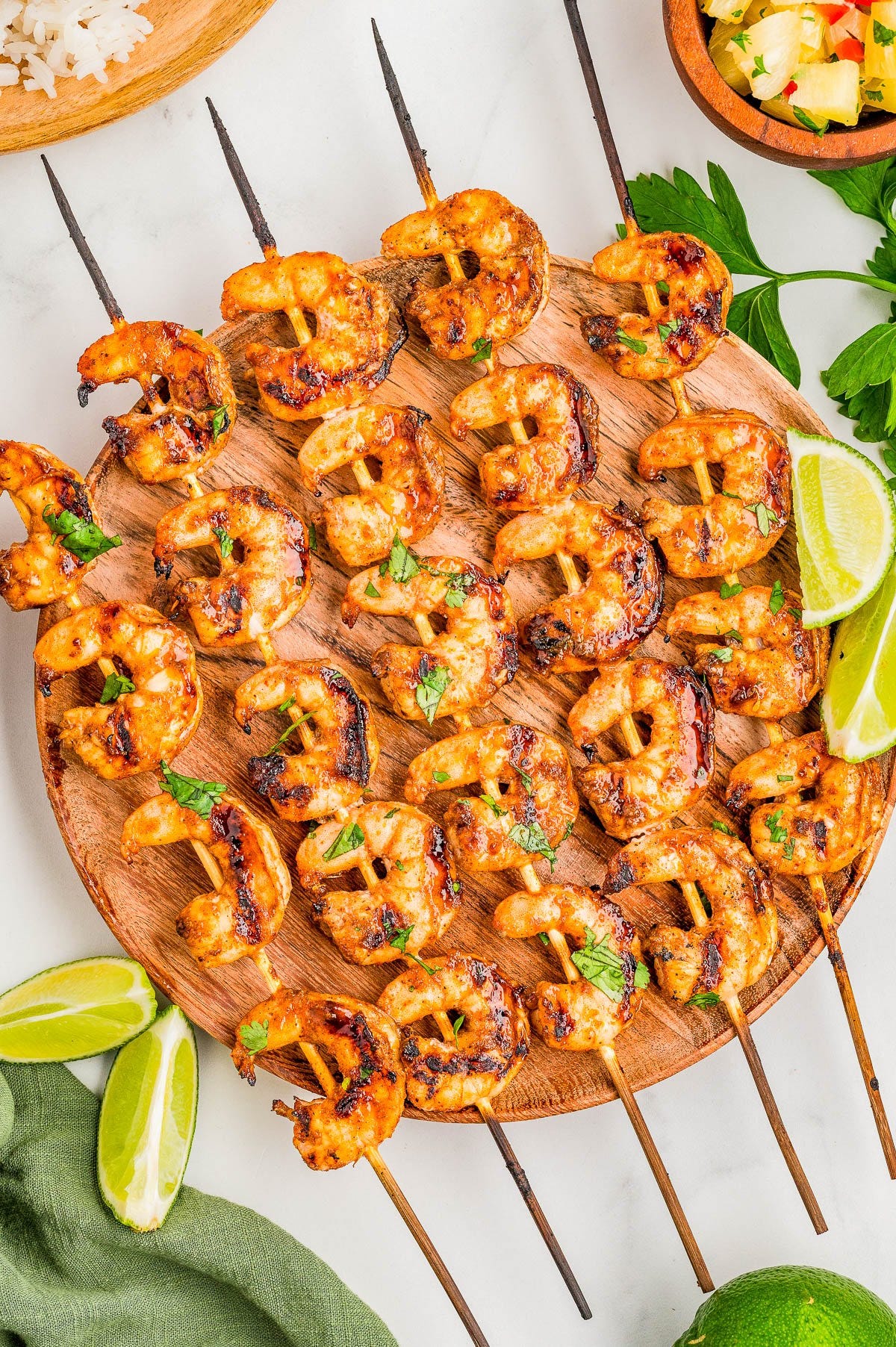 Grilled shrimp skewers arranged on a round wooden platter, garnished with chopped herbs and served with lime wedges.
