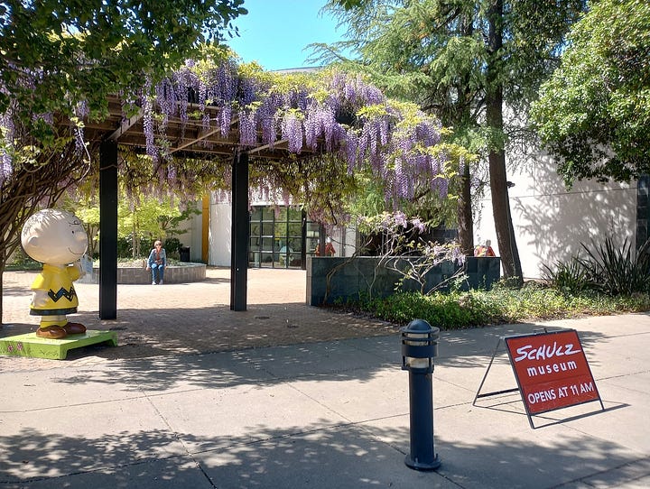 Museum forecourt with Peanuts character statues, trees, flowers, and low entrance doors