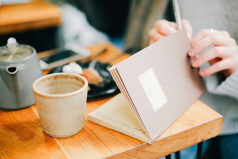 brown table with a mug and pot of tea and plate of food and an iphone on it. a persons hands — left hand holding open a journal and right hand holding a pen brown table with a mug and pot of tea and plate of food and an iphone on it. a persons hands — left hand holding open a journal and right hand holding a pen
