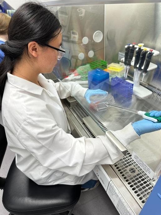 A scientist wearing a white lab coat and blue gloves uses a pipette to work with materials inside a laboratory biosafety cabinet.