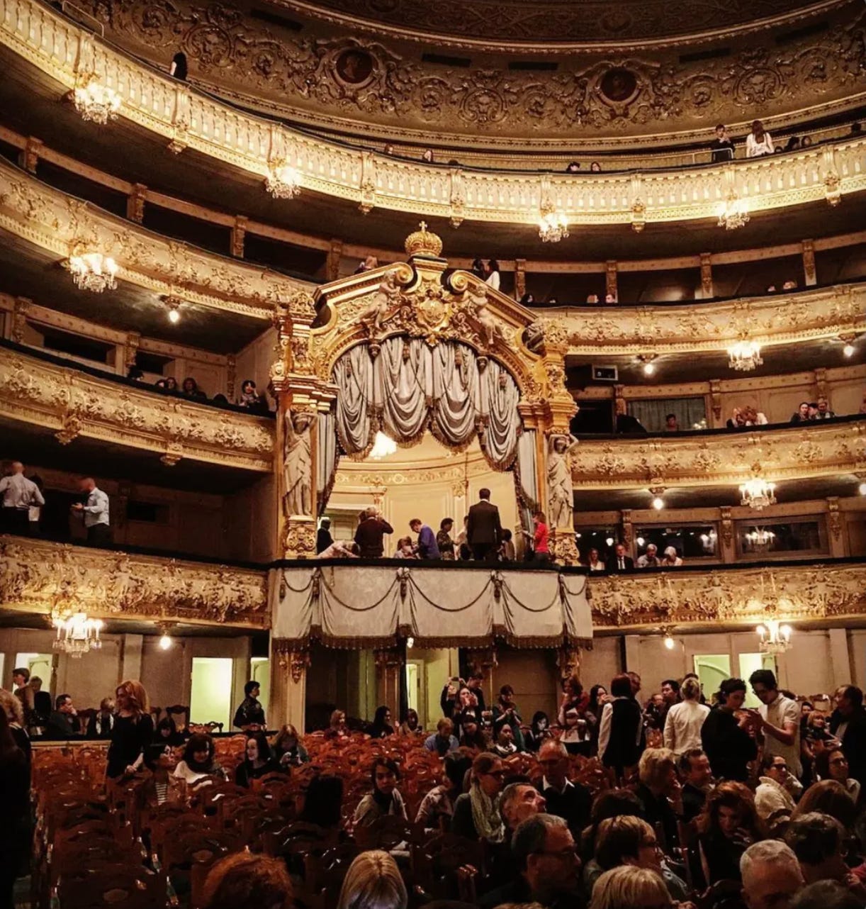 Interior of the Mariinsky Theatre showing multiple gilded balcony tiers, ornate chandeliers, a draped royal box, and crowds gathering on the main floor before a performance.