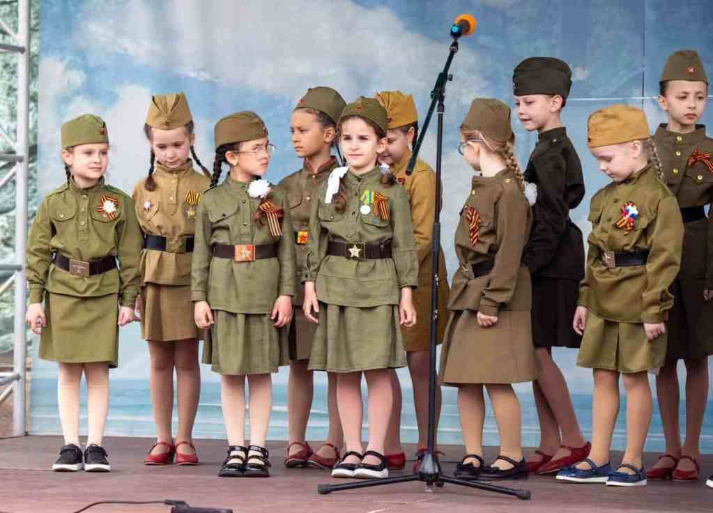 A group of children in military uniforms stand on a stage. The children are wearing green uniforms and are standing in a line