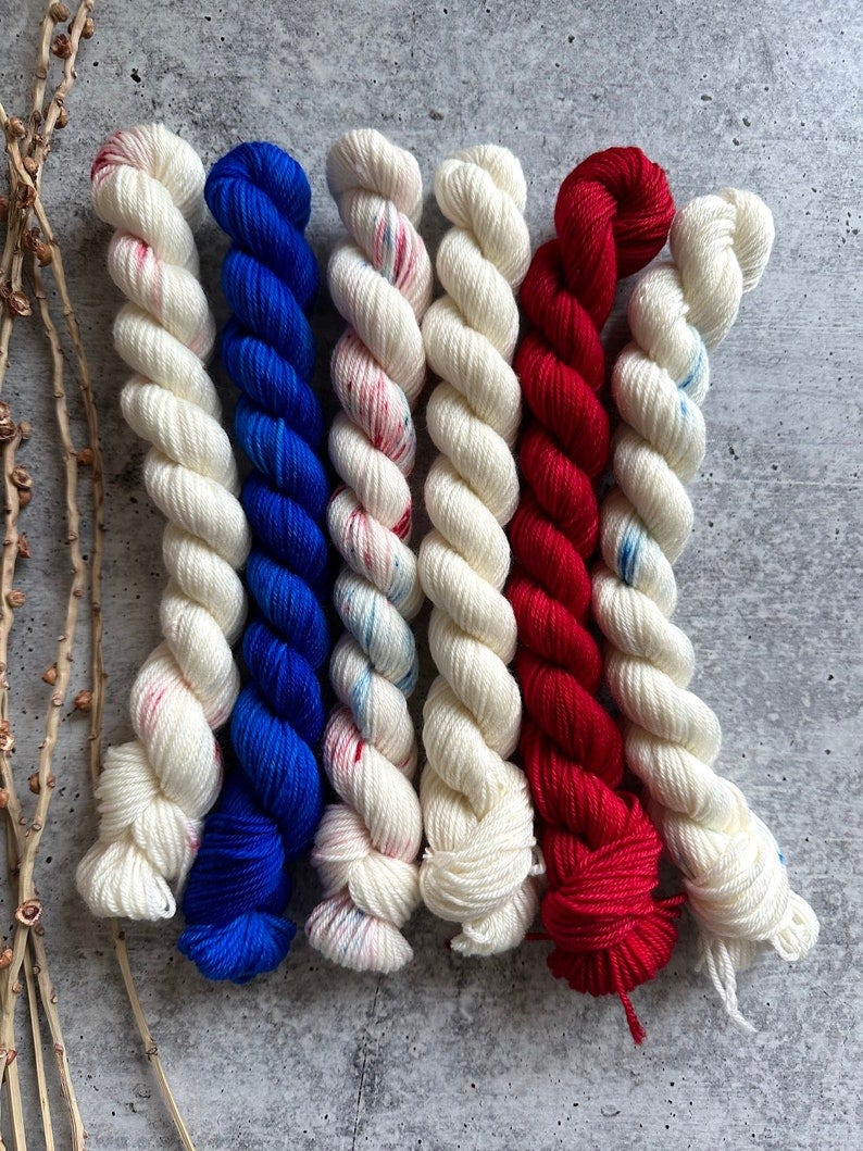 May include: Six skeins of yarn in red, white, and blue colors. The yarn is arranged in a row, with the red skein on the left, the white skein in the middle, and the blue skein on the right. The yarn is twisted and knotted, and the colors are vibrant and bright.