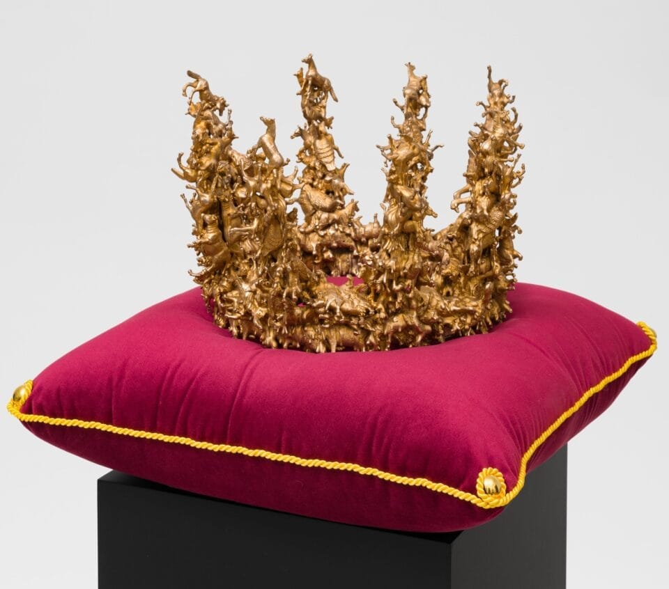 a sculpture of a bronze crown composed of tiny birds and other animals, coated in metallic silver, situated on a red velvet cushion